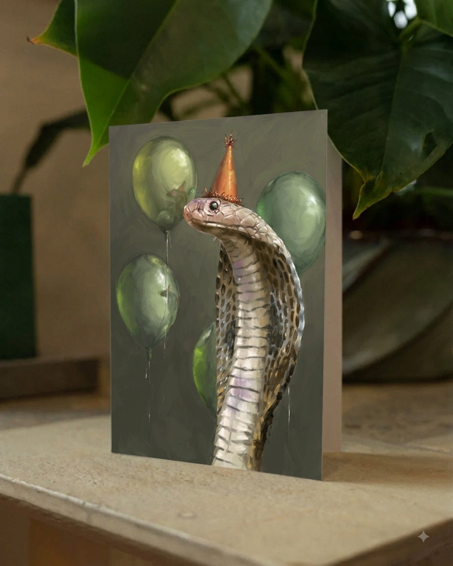 A greeting card featuring a painted cobra snake with a small party hat and green balloons, displayed upright on a tabletop with large green leaves in the background.