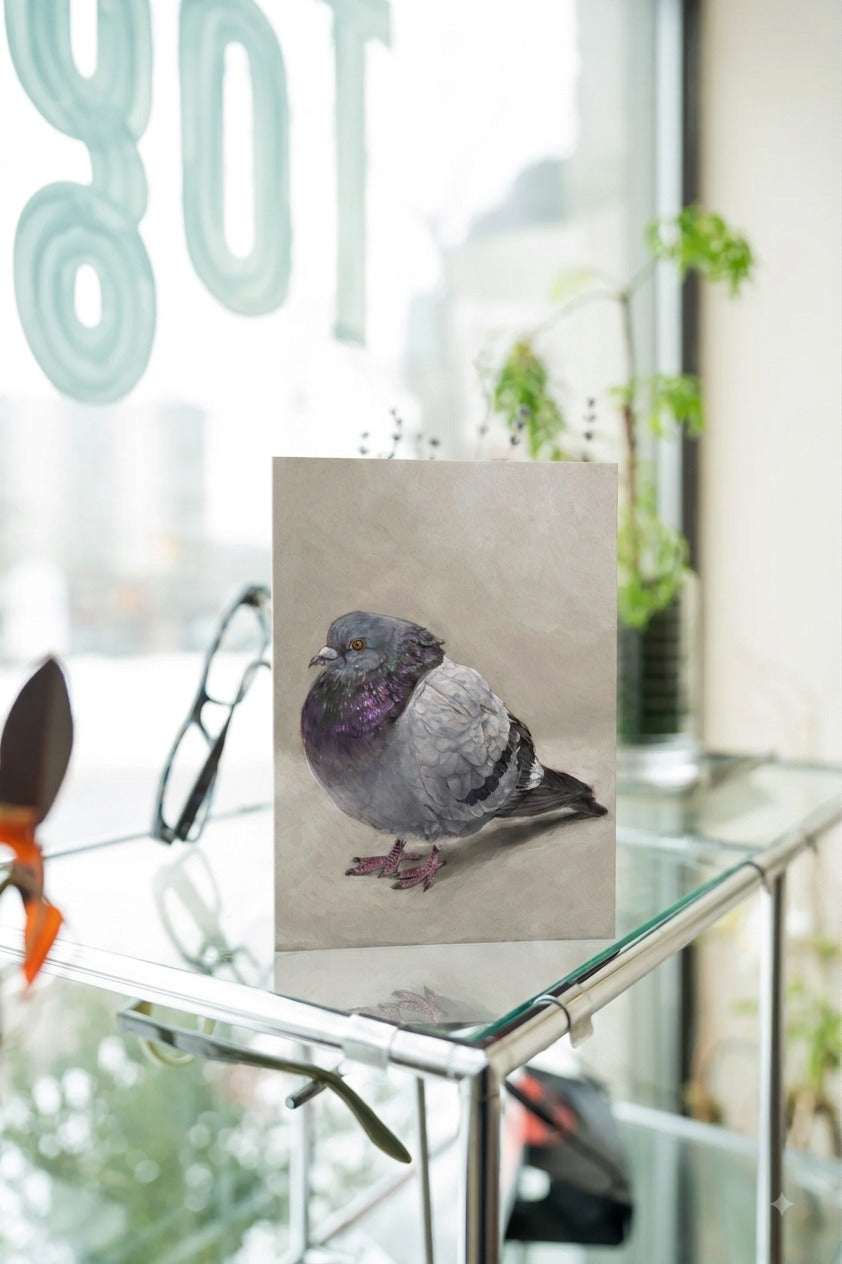 A greeting card featuring a detailed painting of a pigeon with iridescent purple and gray feathers, displayed on a glass table in a bright, modern room.